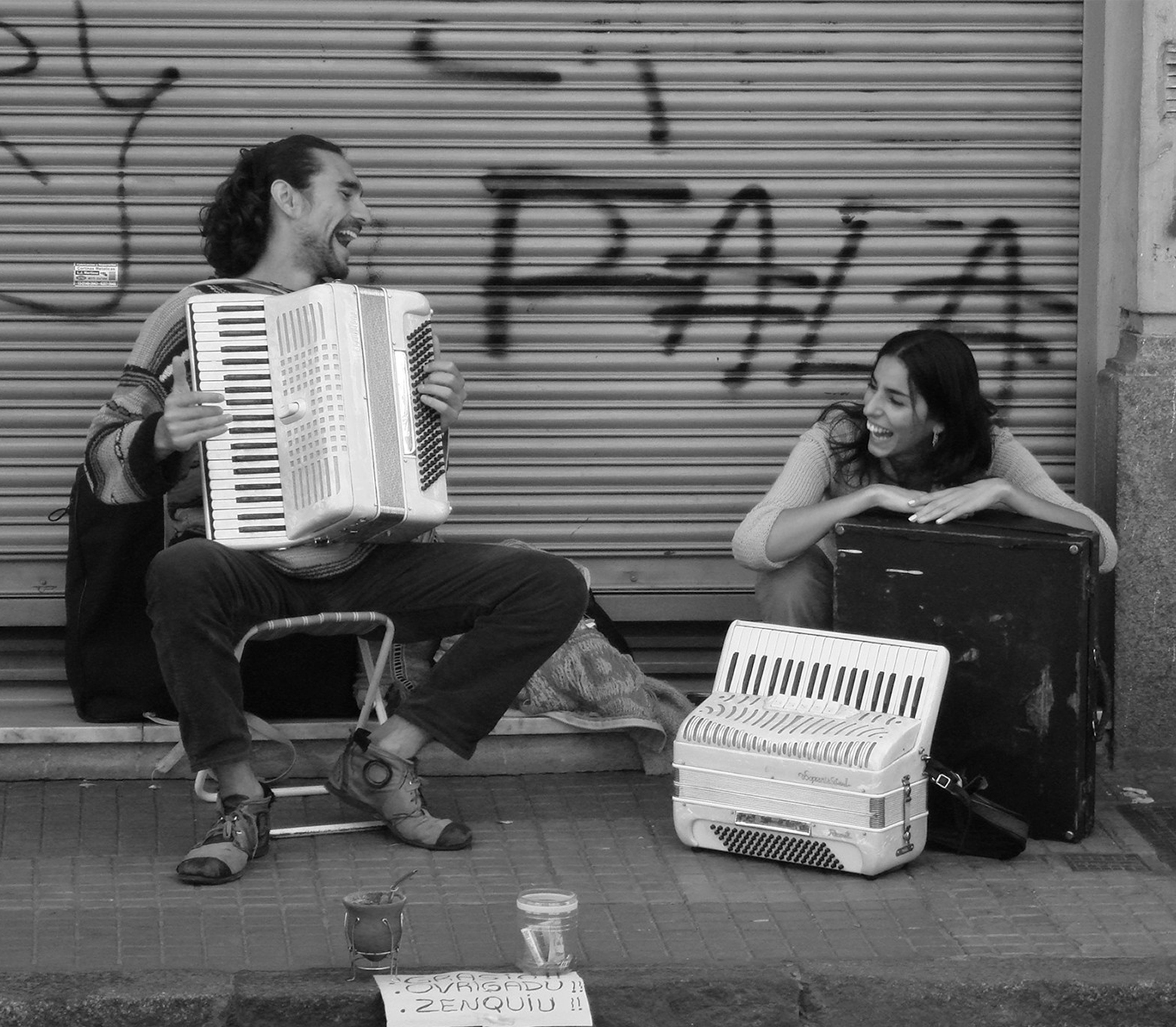 Two accordion players on the street laughing and smiling at each other. The man is sitting on a chair with an accordion on his lap. The woman is crouched down with an accordion on the sidewalk in front of her.